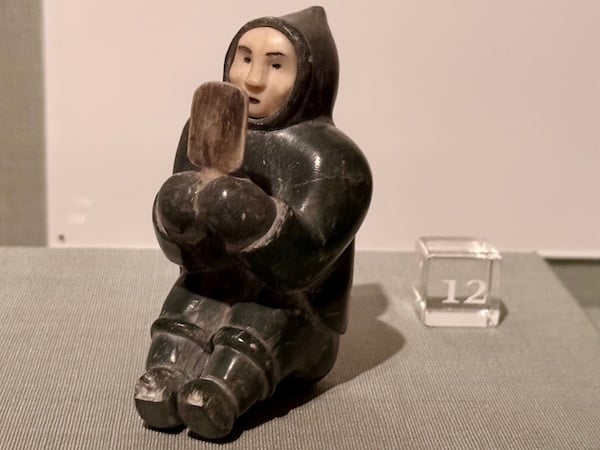 A seated, hooded figure carved from black stone holds and looks into a mirror. The face and mirror of the figure are carved from white ivory.
