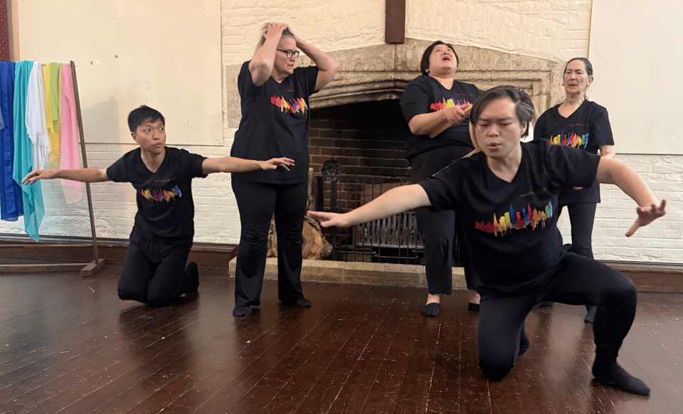 Five performers in black shirts with colorful designs are engaged in a dramatic scene, with one person kneeling and others expressing various emotions, against a backdrop of a rustic fireplace and hanging colorful fabrics.