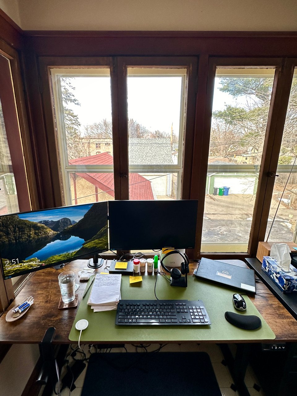 Photo of two computer monitors and other computer accessories on a wood tabletop, with windows overlooking two alleys