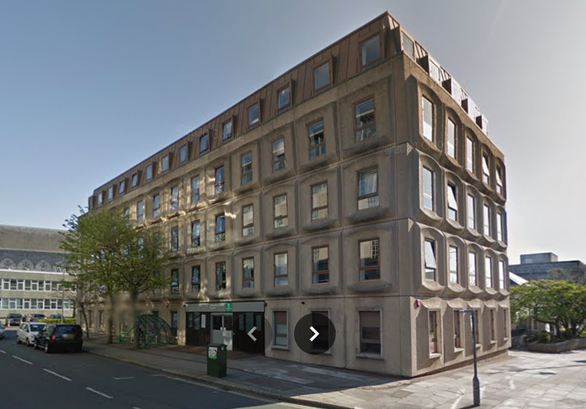 A 1970s style concrete building, seen from one corner. It has four storeys of square concrete panels with windows set in them, and a Mansard roof with more windows. There are a couple of trees near the door on the ground floor. The windows are reflecting the light rather nicely.