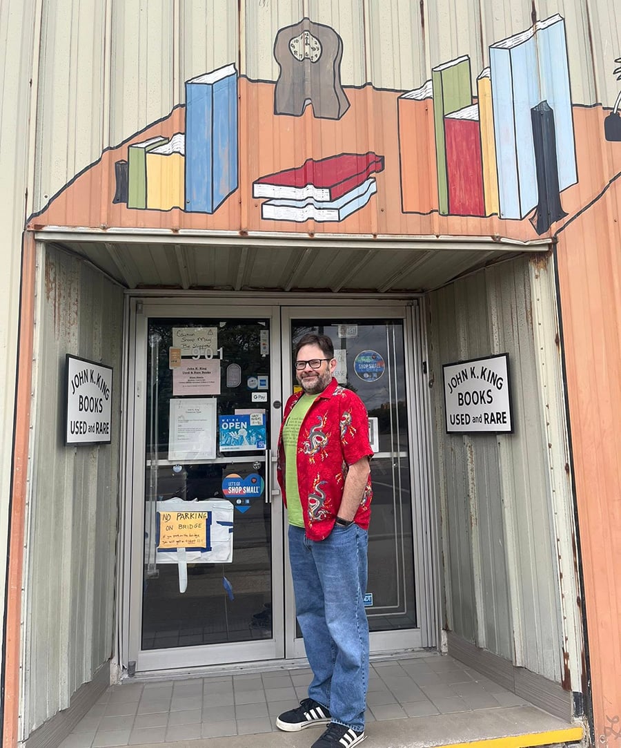 A 50 year old man in a red shirt and cheap jeans stands in front of the entrance to the bookstore.