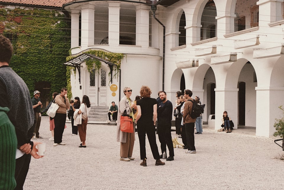 [People standing around and talking in the courtyard of a white castle with arches and ivy.]