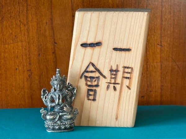 A small silver-colored statue of a multiarmed Buddha figure (Tara) sits on a teal-colored fabric book cover next to a block of balsa wood that has the characters 一期一會 (ichigo ichie) burned into it. The background is cherry-colored wood.