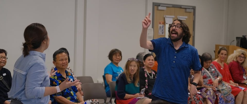 A group of engaged older adults watches a man animatedly speaking while a woman stands facing him, both in a community setting.