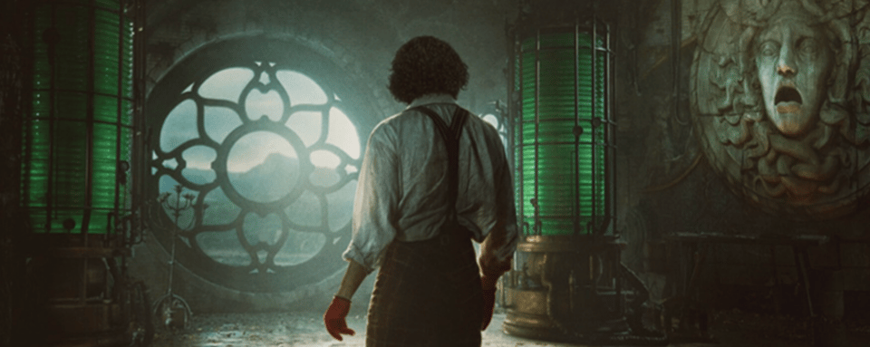 An elaborate gothic lab in hues of green and grey, with a rosette window and a gorgon head on the wall. A man stands with his back to the viewer, his hands red.