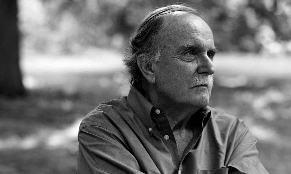 A black-and-white photo of Alvin Lucier. He is outdoors in a park, in the foreground, looking off to the right. He is balding with thinning hair and a thick white mustache. In the background, in soft focus, are grass, a tree and leaves.