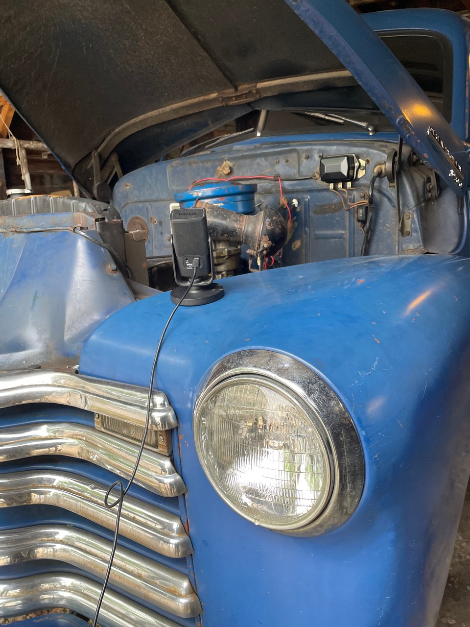 A small microphone sits on a massive blue truck. The truck's hood is open exposing the twisting wires and rusted metal of the engine