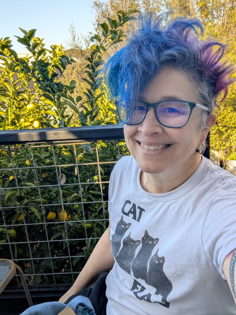 Liz smiling, wearing a tshirt that says CAT FLAG with black cats in the shape of the logo of the punk band Black Flag. Lemon tree and blue sky in the background.