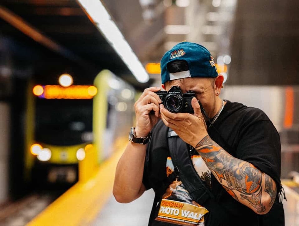 Picture of me holding a black 35mm film camera (namely my Pentax MX, the first camera I ever owned), wearing a black t-shirt with a design reading "Crenshaw Photo Walk," in an underground L.A. Metro station as a Metro train comes into the frame.