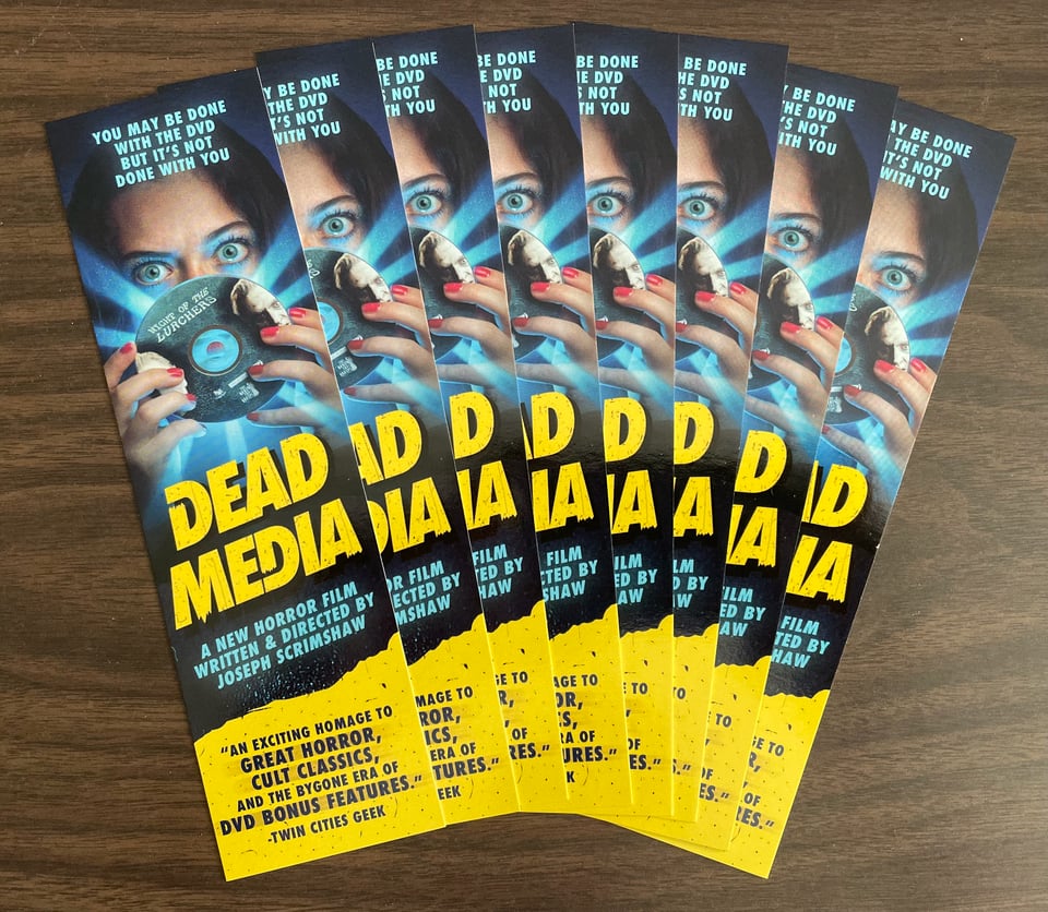 Promotional cards for the horror film DEAD MEDIA fanned out on a table. The card features a woman peering over a glowing DVD.