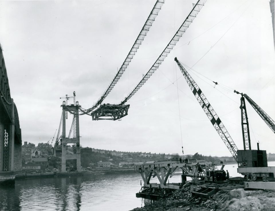 The suspension bridge under construction. Metal cables have been slung between the concrete towers and the first section of roadway is hanging off them.