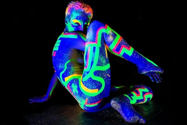 En una escena a oscuras, una modelo desnuda y con pelo corto con pinturas brillantes de neón describiendo patrones geométricos sobre su cuerpo.