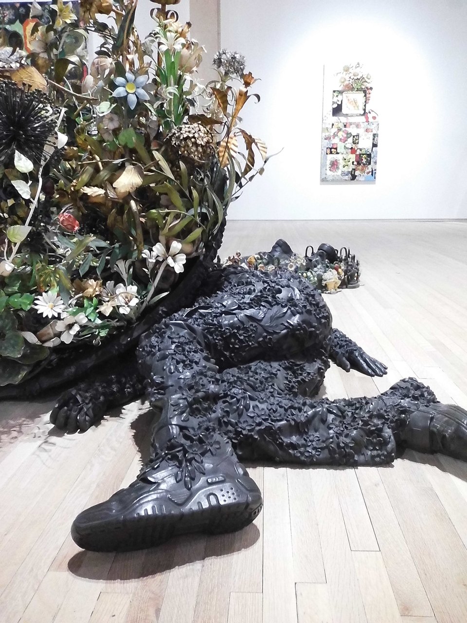 Digital instax photo of part of a Nick Cave sculpture, a bronze body lying on its side, composed of flora, beside a large artificial flower arrangement