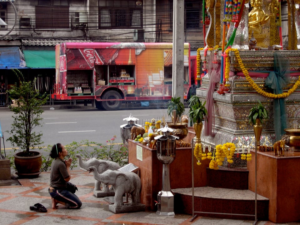 In the foreground a woman kneels in front of a gold statue decorated with flowers, praying. In the background we see an empty road and old grey buildings. The only vehicle is a large, red, Coca-Cola truck.