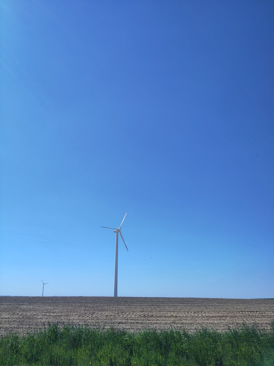 Brachfläche, im Vordergrund etwas Gras. Im Hintergrund ragen zwei Windräder in die Höhe.