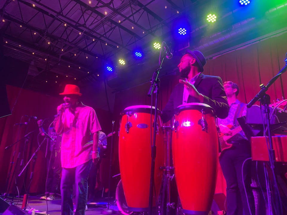 A band on stage, one person singing, another playing the congas, another on bass