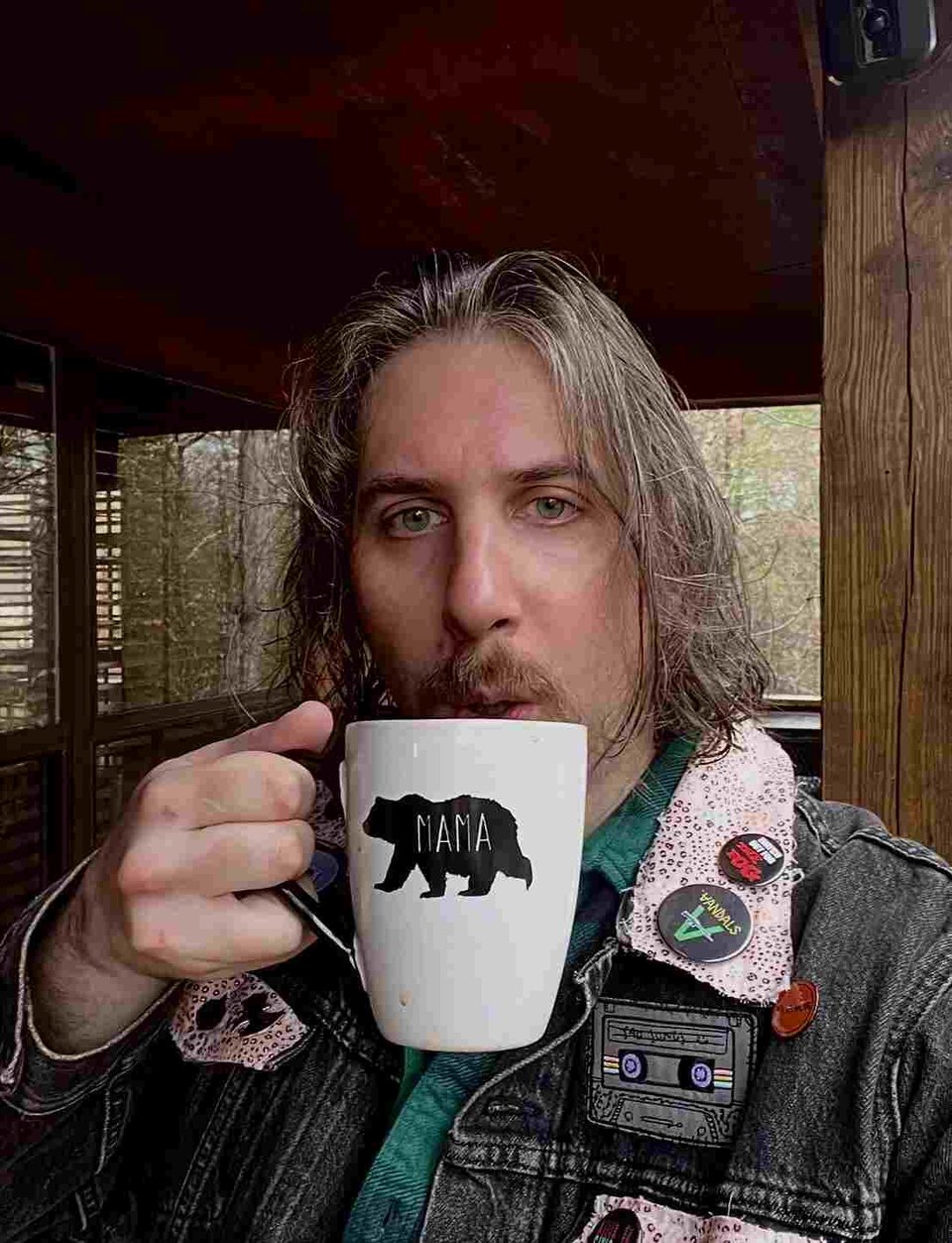 A person with chin-length hair and a mustache wears a gray denim jacket with patches and holds up in their right hand a white coffee mug with the silhouette of a bear that reads "MAMA"