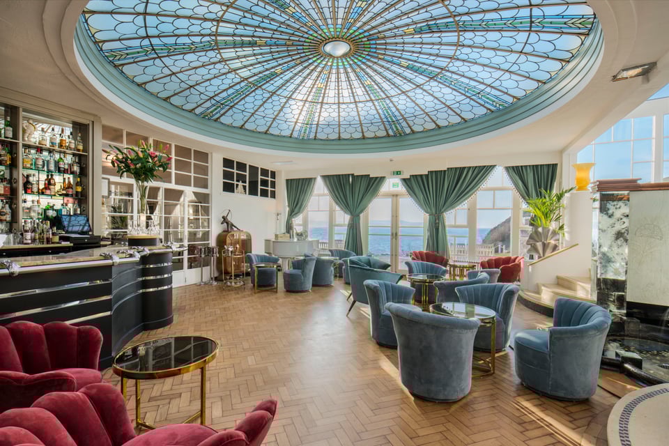 A high res photo of the cocktail lounge under the peacock dome. The dome is made up of small panes of pastel and clear glass cut to resemble stylised overlapping feathers. On the herringbone parquet floor, clusters of velvet bucket chairs are scattered, and beyond the wall of windows is the sea.