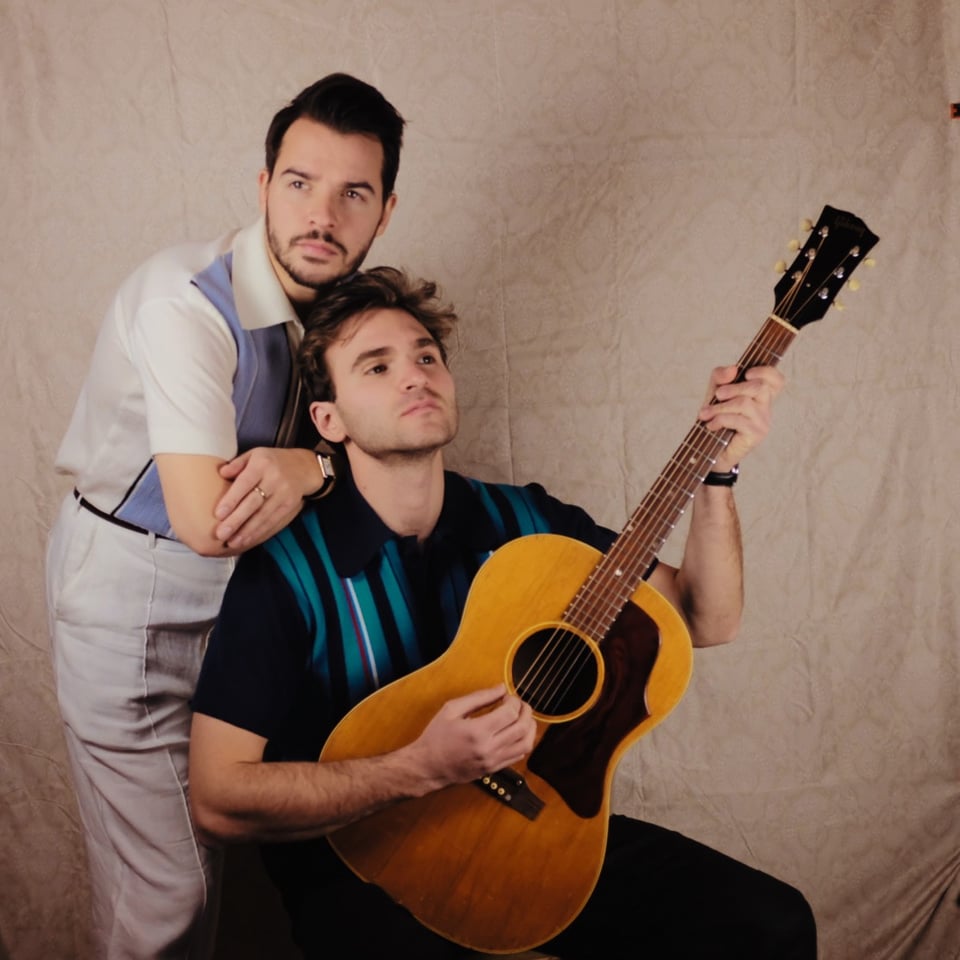 A man with dark hair and trimmed beard, wearing a white and blue print shirt and light pants, stands leaning with his arms on the shoulders of a man in a dark mod-patterned shirt who is sitting and holding an acoustic guitar.