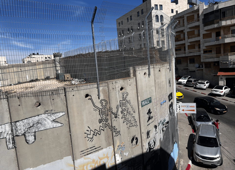 A photograph of a street with a tall concrete wall, composed of separate individual panels. Above the wall is dense wire.  The gray wall is covered with graffiti and small signs (including one that says 'Wall St').  There are parked cars, an apartment building, and a blue sky.
