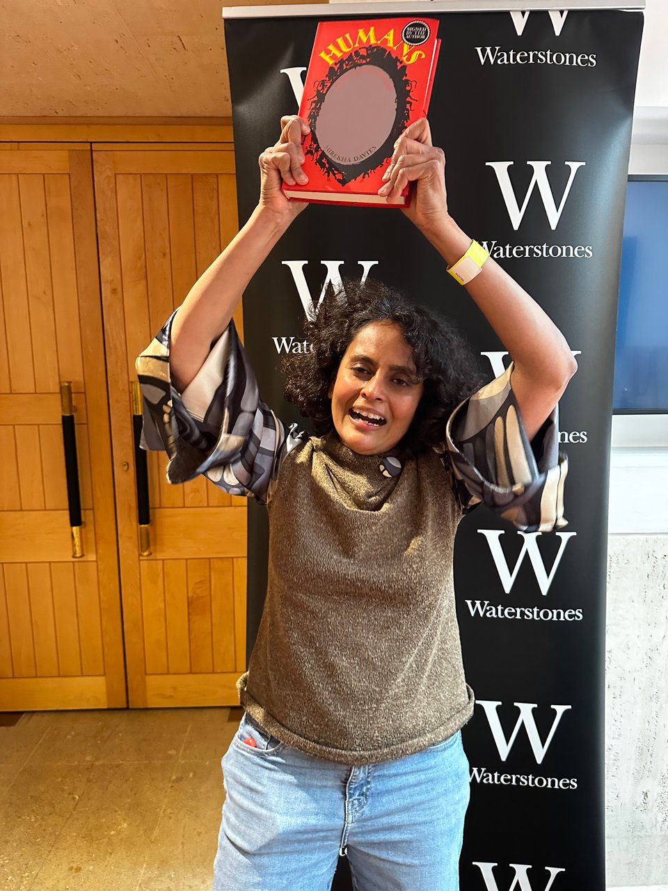 Woman with short, dark hair, in a beige woollen top with blue and beige sleeves holds a bright red book with an oval mirror on the cover up above her head with an air of jubilation. Behind her is a black banner with the Waterstones name and W logo in white. In the lefthand background is a pair of wooden doors.