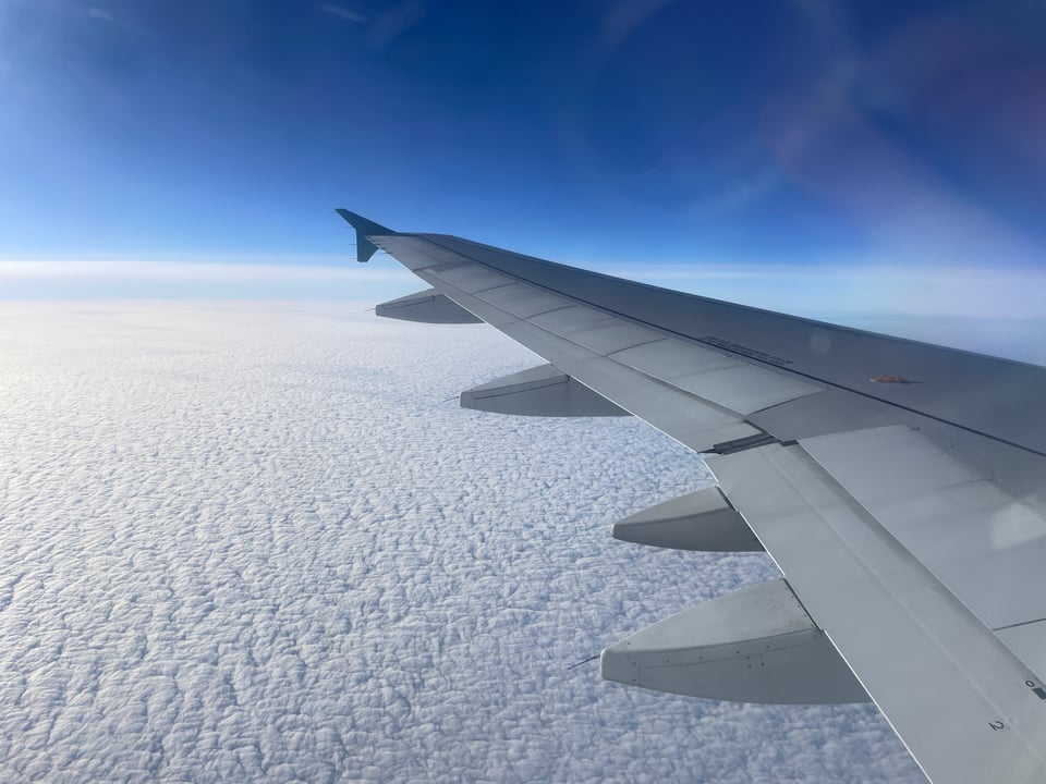 An airplane wing flies over white clouds that cover the earth below. The blue sky is intense above the clouds.