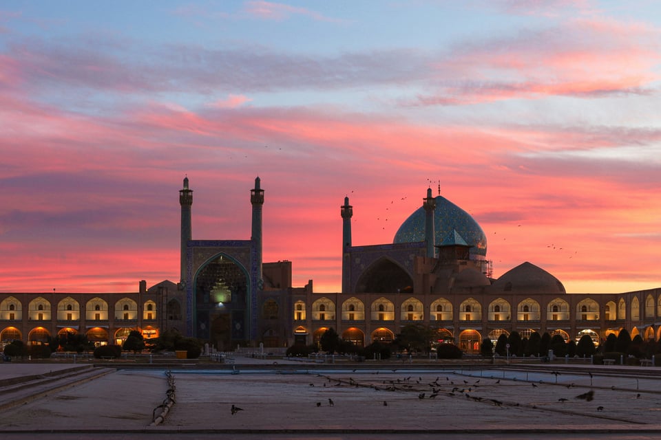 Sunset over the Sheikh Lotfollah Mosque in Iran. The setting sun is painting the broken clouds over the mosque with pink, purple, and orange hues against the blue sky. The mosque's intricate arches are lit as night falls.