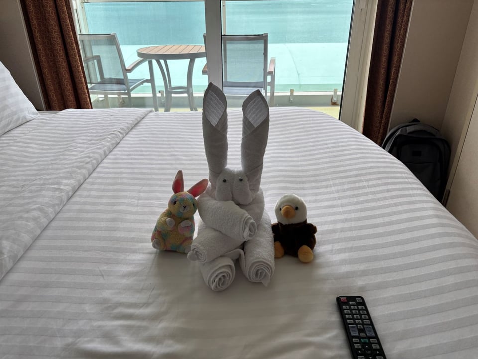 Cruise ship stateroom with a towel bunny with googly eyes sitting on the made bed, in between a small stuffed rainbow hued bunny and a stuffy. The towel bunny's arms are crossed as if in impatience.