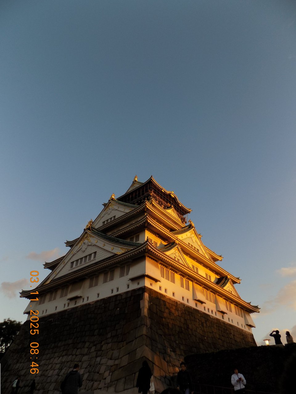 A photograph of Osaka castle at sunset, which illuminates the massive building in a golden light. The sky is mostly clear of clouds. The photo is taken from close to the bottom of the building, pointing up. Some other tourists can be seen taking photographs.