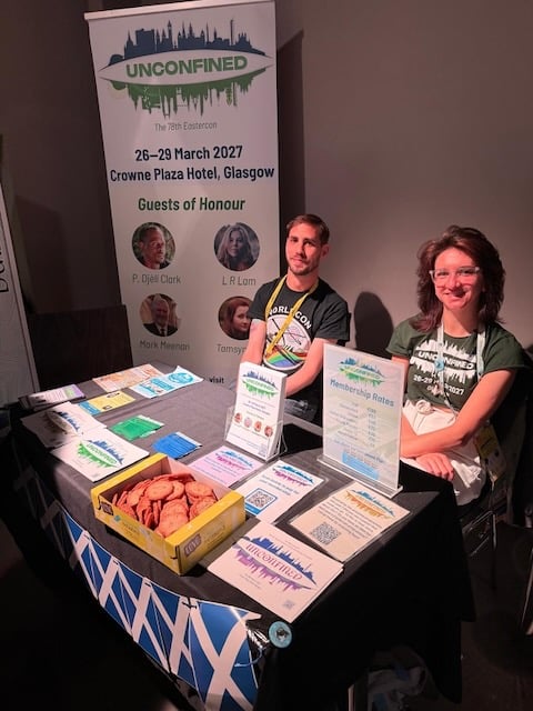 Two people sat behind a table covered in leaflets and ribbons promoting the convention, plus biscuits.