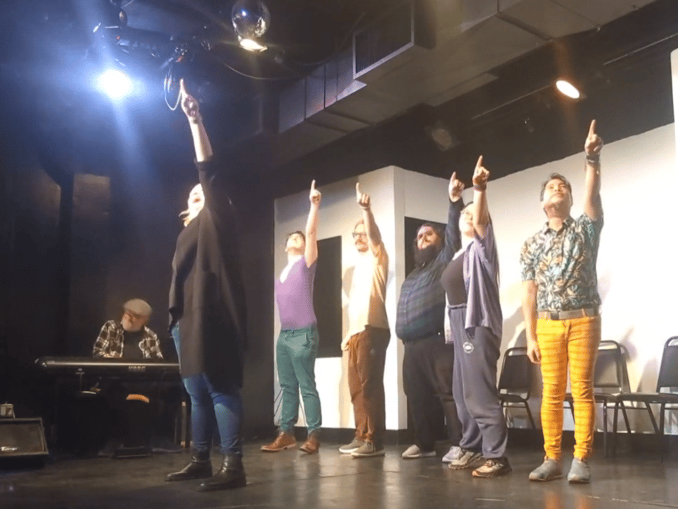 A moment in a musical improv show: Six improvisers on stage, pointing up at something in space. A piano player in the background.