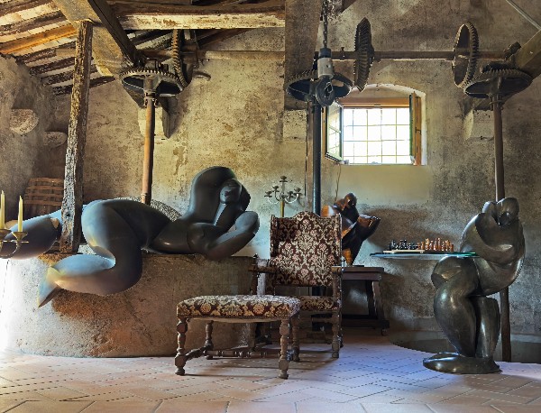 Stone-walled room with exposed wooden beams, an old olive press, and abstract human sculptures.