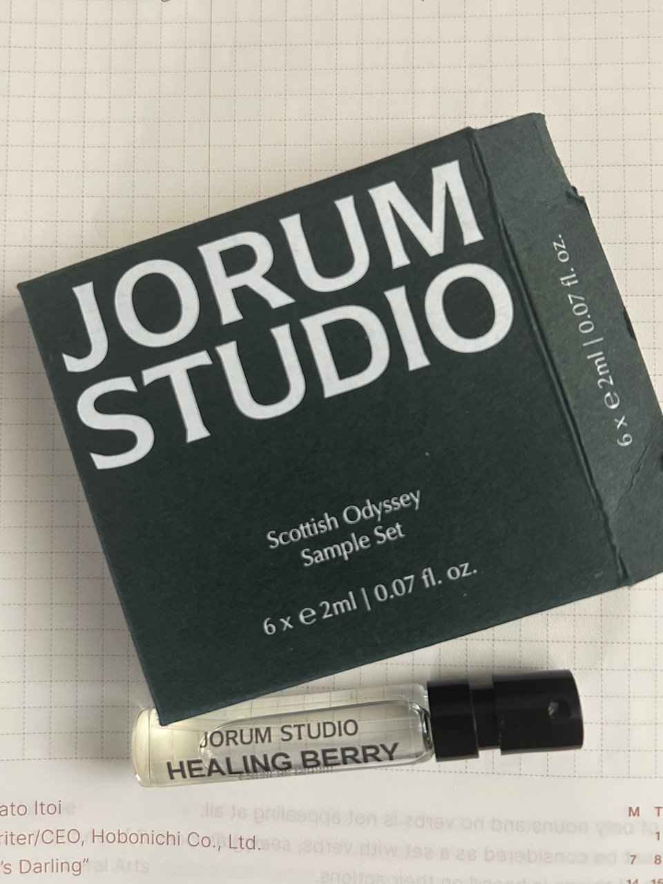 A small glass vial of perfume is next to the dark green Jorum Studio box. Everything is on a squared paper notebook.