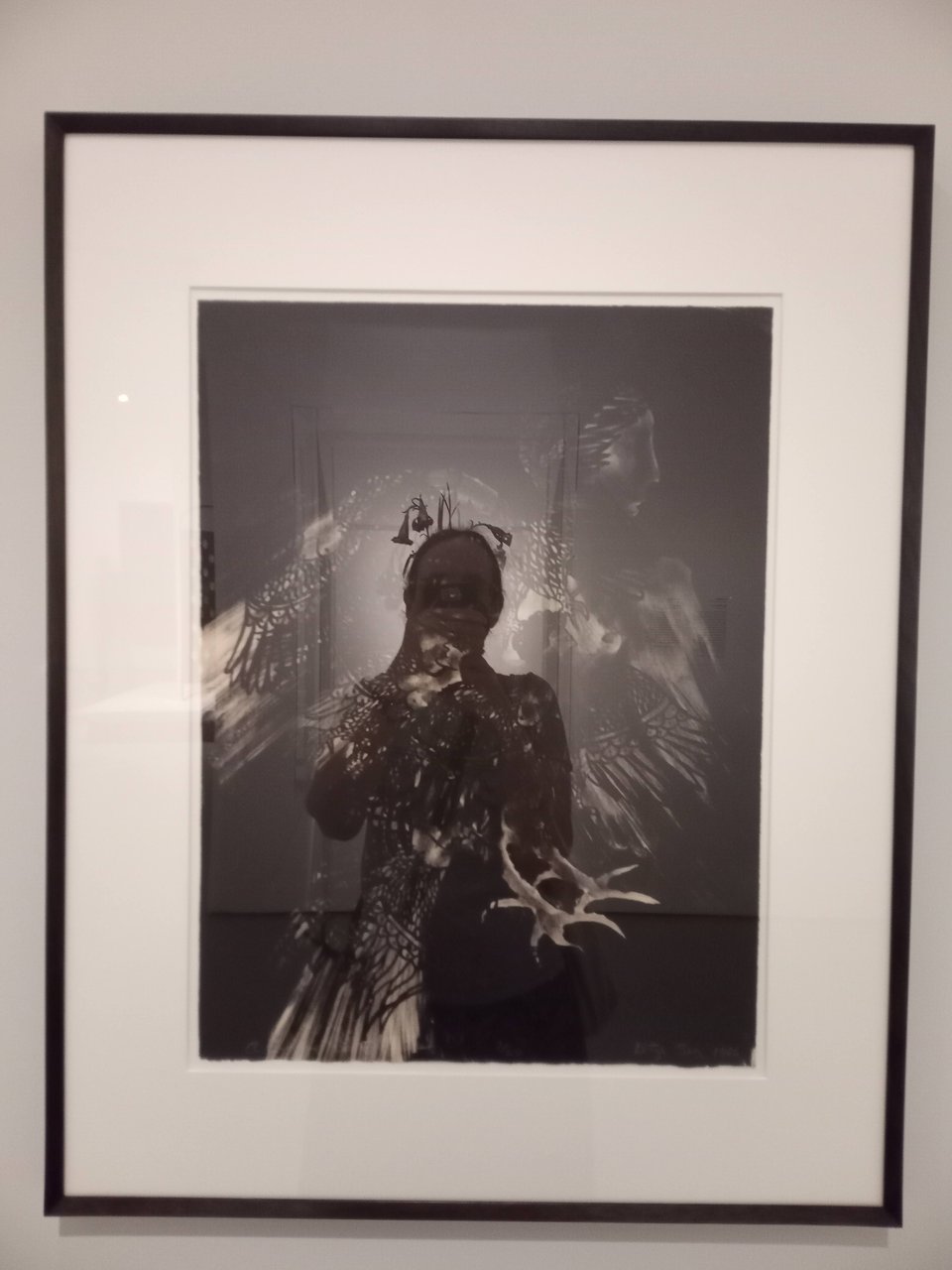 A photo of "A Siege of Sirens" by Betye Saar, which is a black and white print of a human-headed bird creature. You can't really see it, because my reflection as I take the photo dominates it. I'm lit from behind like I have a halo or something.