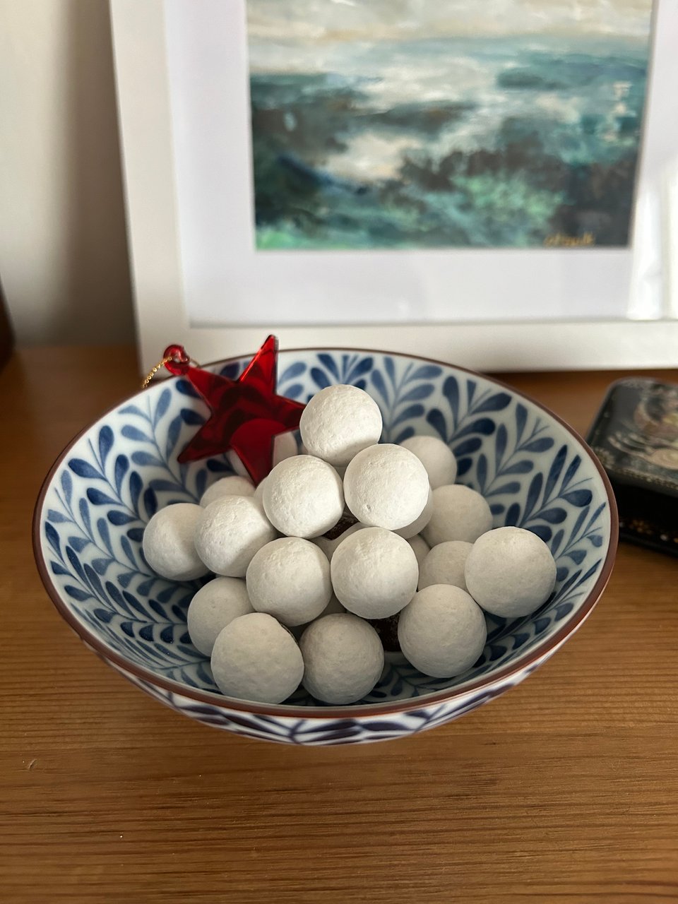 A blue and white Japanese bowl filled with white dusted Cadbury's chocolate snowballs. There is a red glass star perched in the bowl and a small seascape behind.