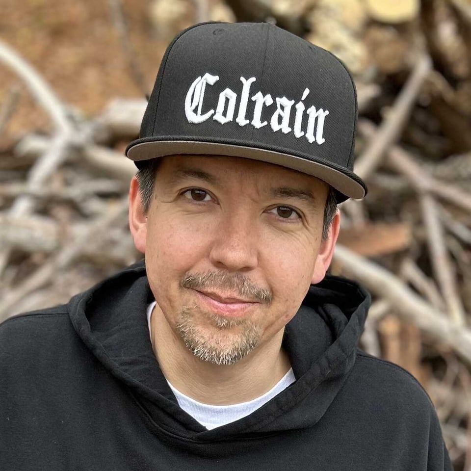 A close-up of a man with a short-clipped goatee wearing a black baseball cap with the word "COLRAIN" in stylized white letters. The neck of a white t-shirt is visible under a black hoodie.
