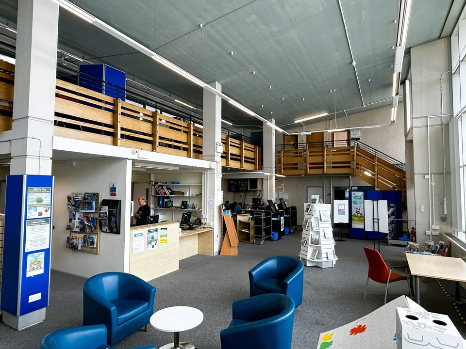 An internal photo of the original building from the north west corner. H shaped beams hold up the student gallery, as before. The centre of the library has been boxed in to create a reception area and office. Bucket chairs are arranged around a low table. The lights are now a long track of fluorescent or LED tubes.