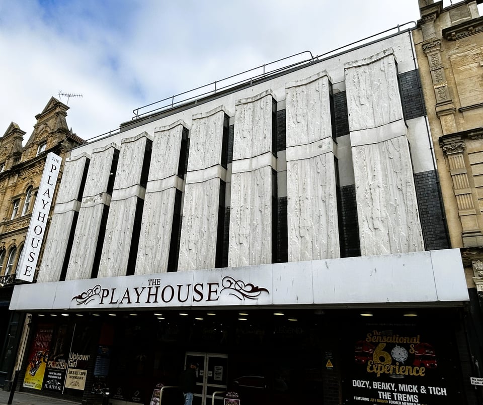A theatre front. At the ground floor, the 1960s wood and glass is barely visible. Above the marquee, the front has eight boxed-out panels rising to two storeys. They are fronted with white fibre-glass molded to look like rough concrete.