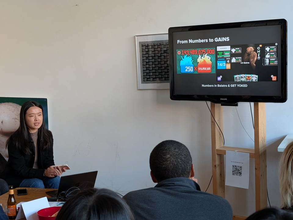 An audience listens to a speaker seated in front a a laptop, with a TV screen behind her. Screen title reads "From Numbers to GAINS"
