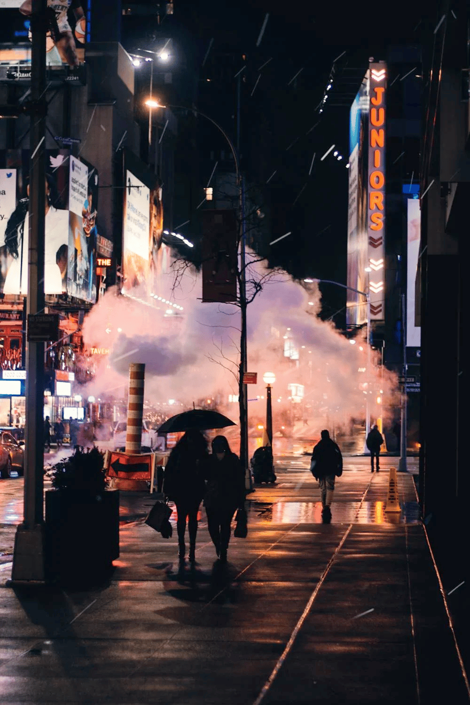 Nighttime street scene in New York City, illuminated by neon signs and glowing storefronts. The "JUNIORS" sign glows in orange letters on the right side, hinting at the location near Times Square. A cloud of steam billows from a street vent, casting a dramatic pinkish hue across the scene. Pedestrians walk along the wet sidewalk, some holding umbrellas, their reflections shimmering on the pavement. Advertisements and billboards tower above, adding to the city's vibrant, cinematic atmosphere. The sidewalks have wet areas reflecting light from above.