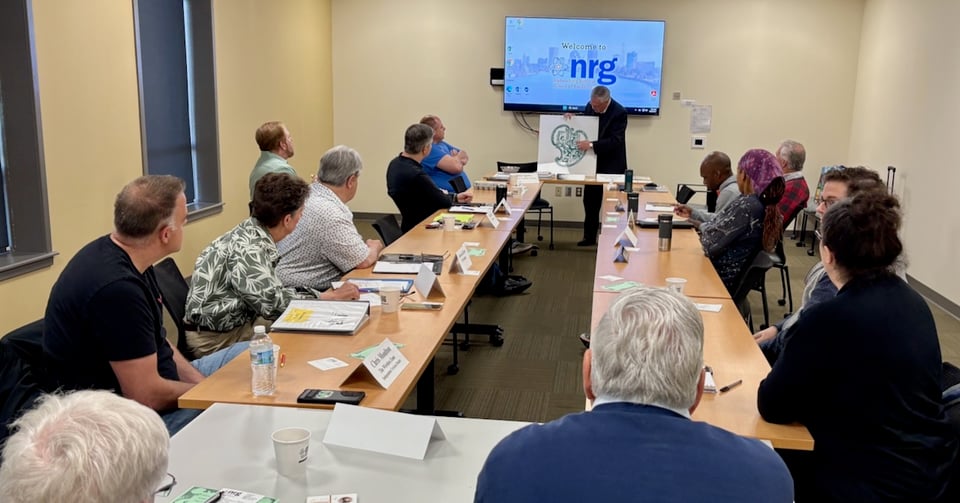 Darryl is at the front of a room of seated people, pointing to the details of a printed logo he’s holding.