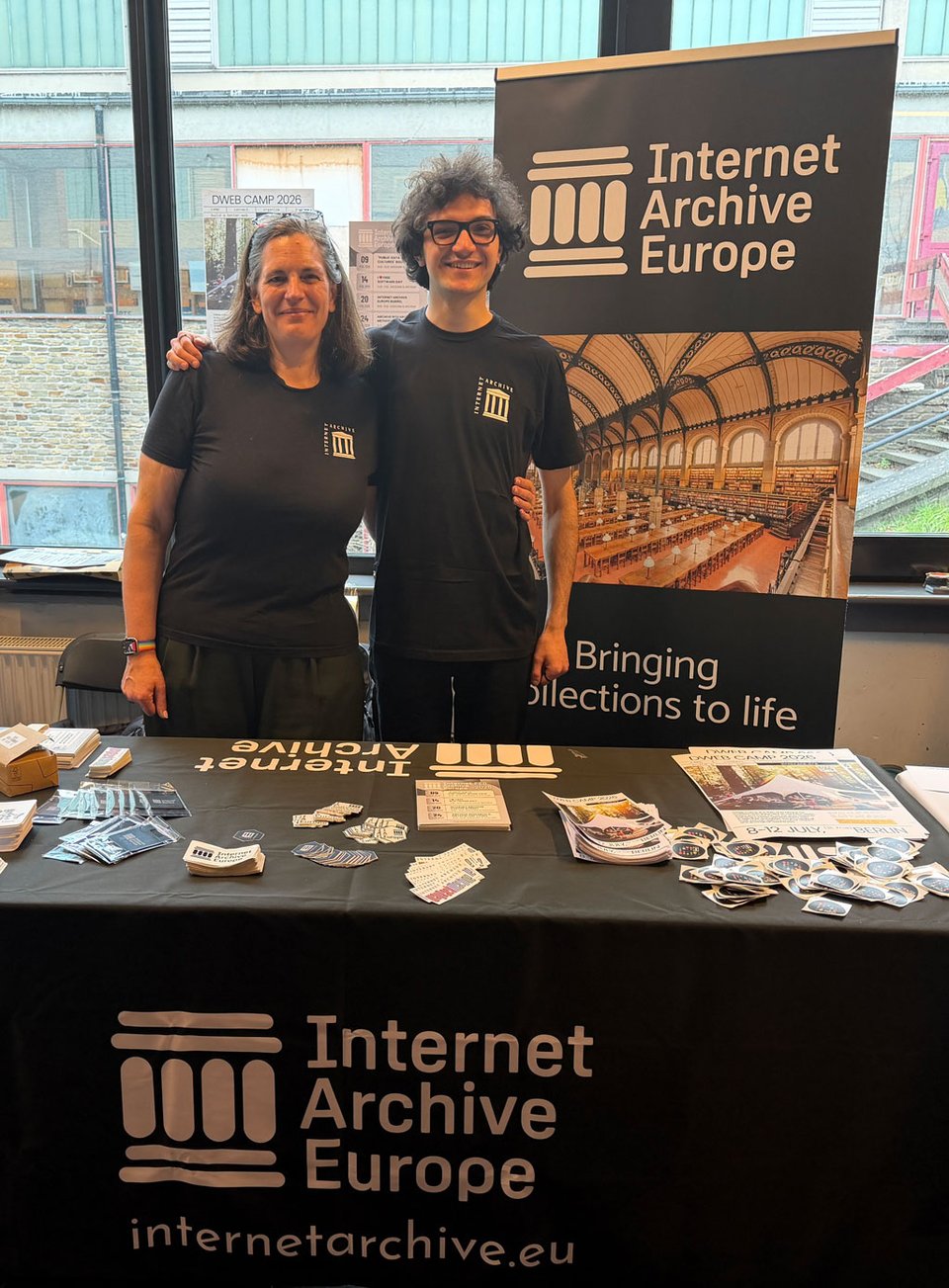 Two people stand behind a table covered with "Internet Archive Europe" stickers and flyers.