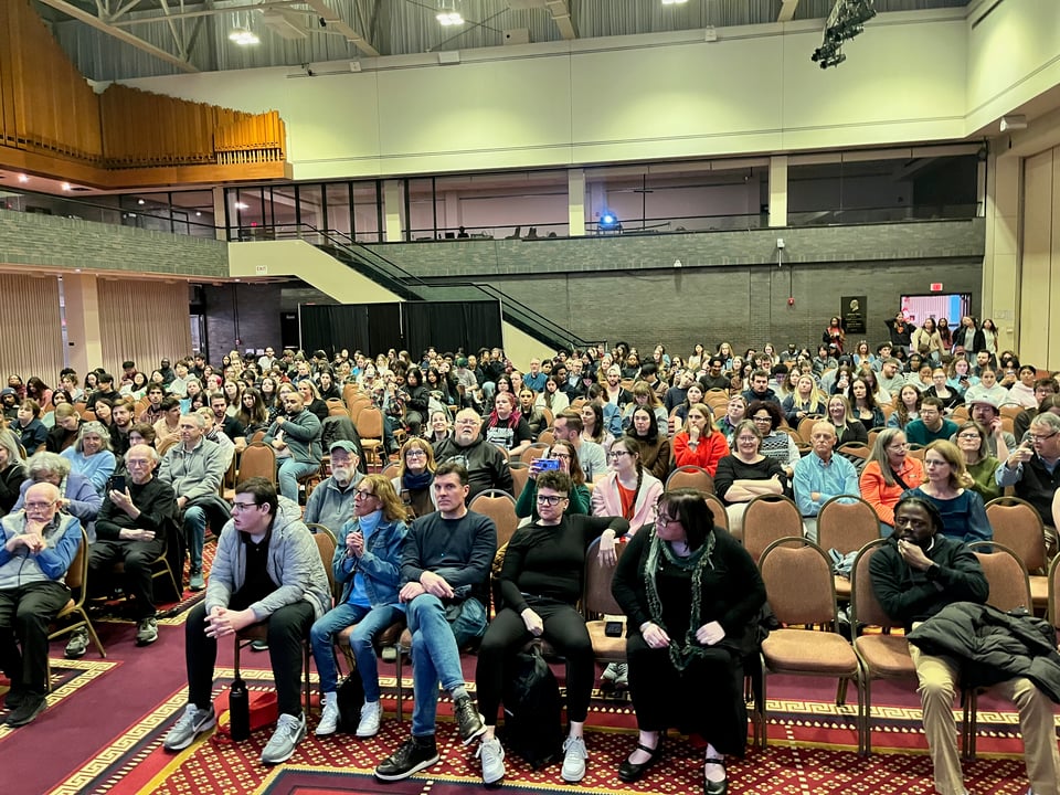 A large crowd (~500 people) sits on chairs in a carpeted college auditorium. The crowd ranges in age from college students up to gray-headed elders.