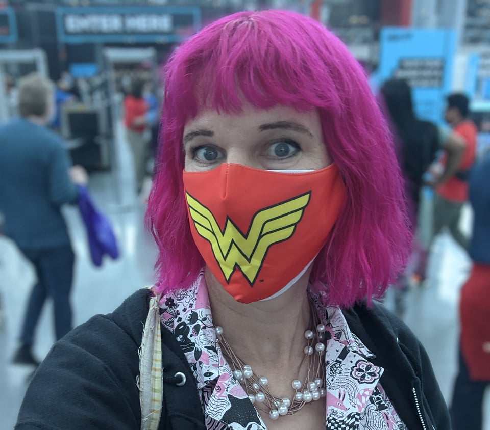 Me at a random convention a while back. I have pink hair in a bob with a fringe, and I'm wearing a Wonder Woman mask over a regular N-95. I also have a necklace with fake pearls, a cute Nooworks pink and white dress, and a black hoodie. The background is blurred.