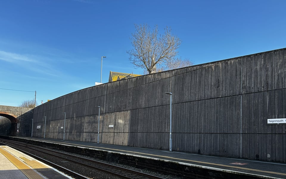 A huge concrete retaining wall behind a long railway platform. The wall is made up of large panels of shuttered concrete and curves up to perhaps 20 foot high. A large section of it is less discloured, suggesting the large advertising hoardings that used to be there.