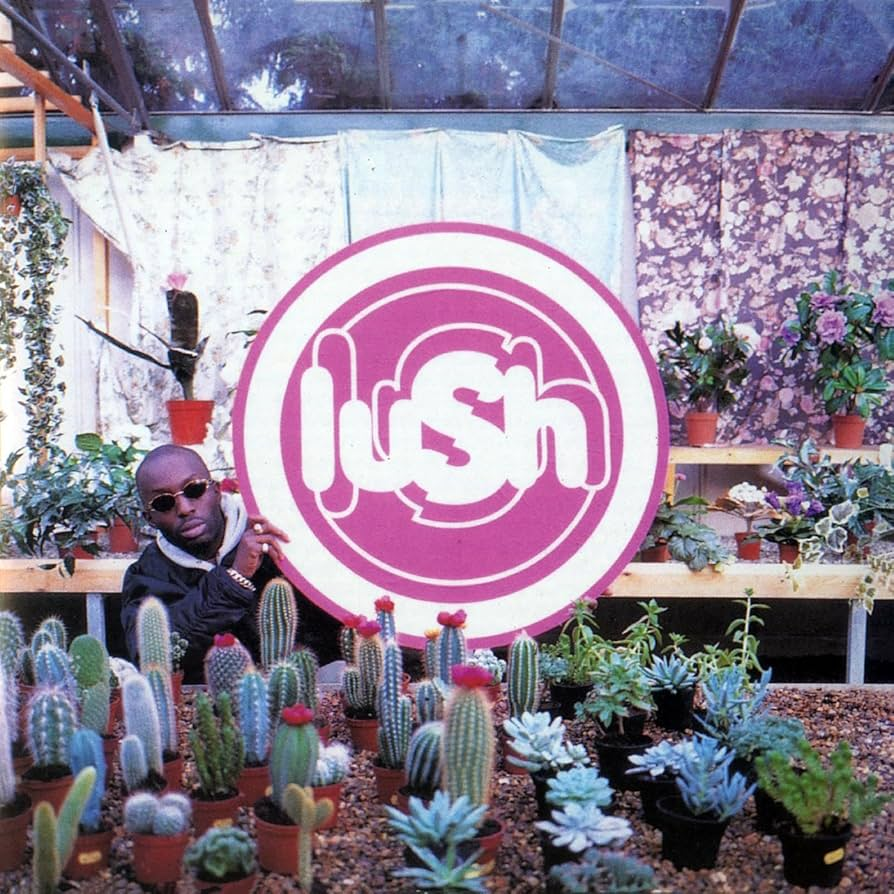 A man in sunglasses holds up a Warhol like Pop sign saying Lush, in what appears to be a florists.
