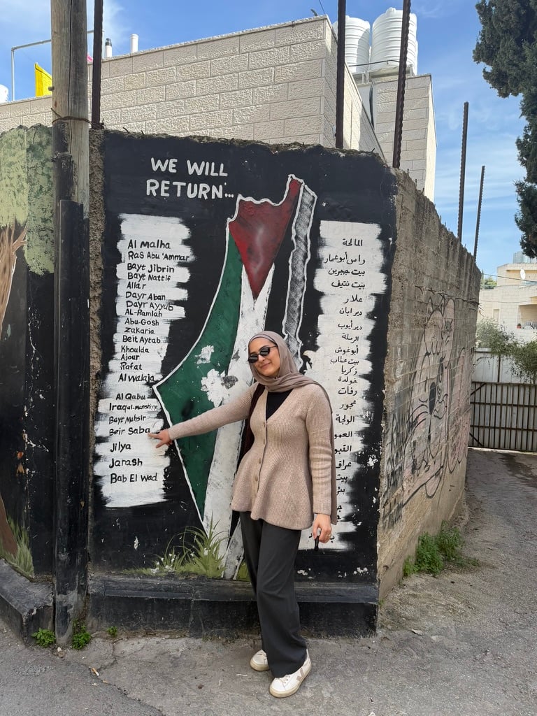 Photograph of a woman standing in front of a mural featuring a map of Palestine colored with the Palestinian flag and the phrase "WE WILL RETURN..." above it. The mural lists names of various locations in Arabic and English on either side of the map. In the background is a cinderblock building with large white tanks atop the roof.