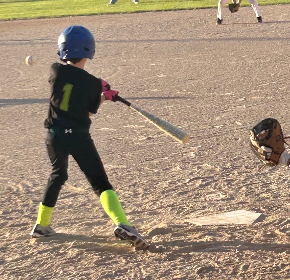 The back of a little league baseball player in a black uniform and blue helmet, swinging at a pitch. You can see the baseball in the air.