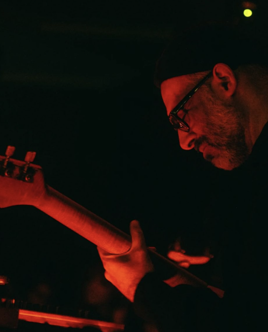 Dirar Kalash, bathed in orange light, wearing black beanie, black hoodie and spectacles, plays a bass guitar