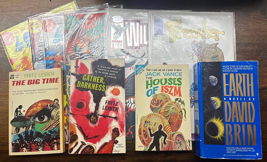 Picture shows some comic books in plastic; the way the light is hitting the plastic they’re hard to se, but they’re three issues of Legion of Super-Heroes, a Green Arrow, an issue of Wildstar, and an issue of Trencher. on top of the comics are four paperbacks—The Big Time, Gather Darkness, Houses of Iszm, and Earth, all science fiction.